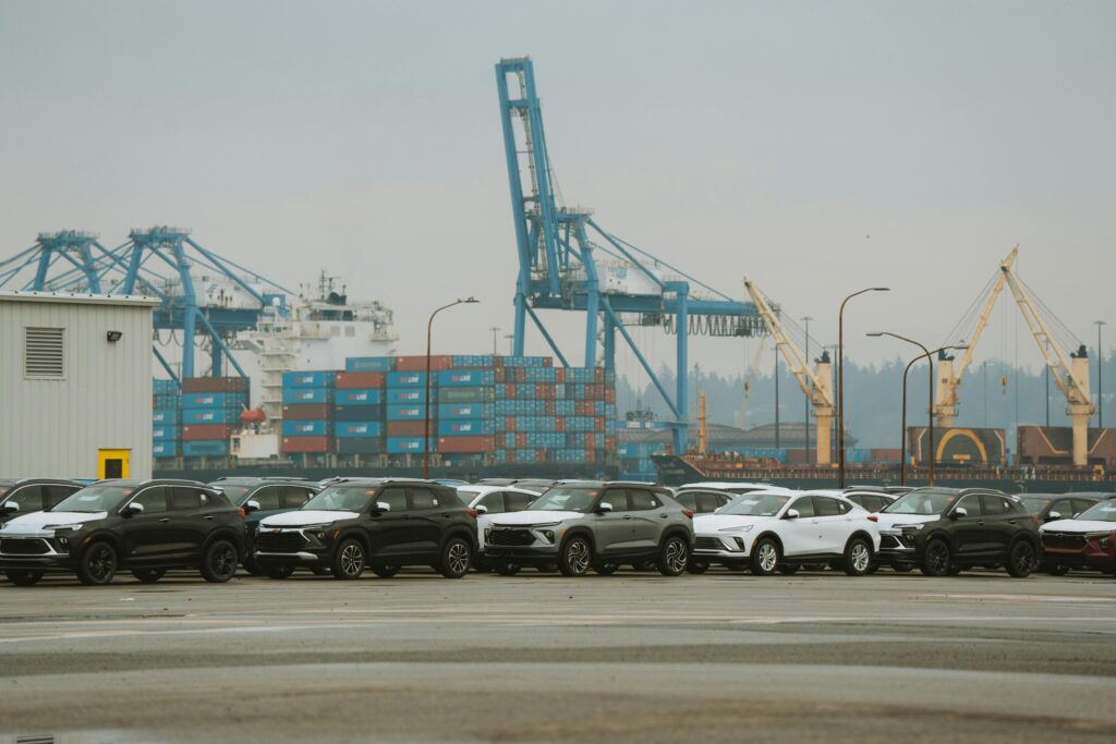 A lineup of parked cars at a bustling industrial shipping port with cranes and containers.